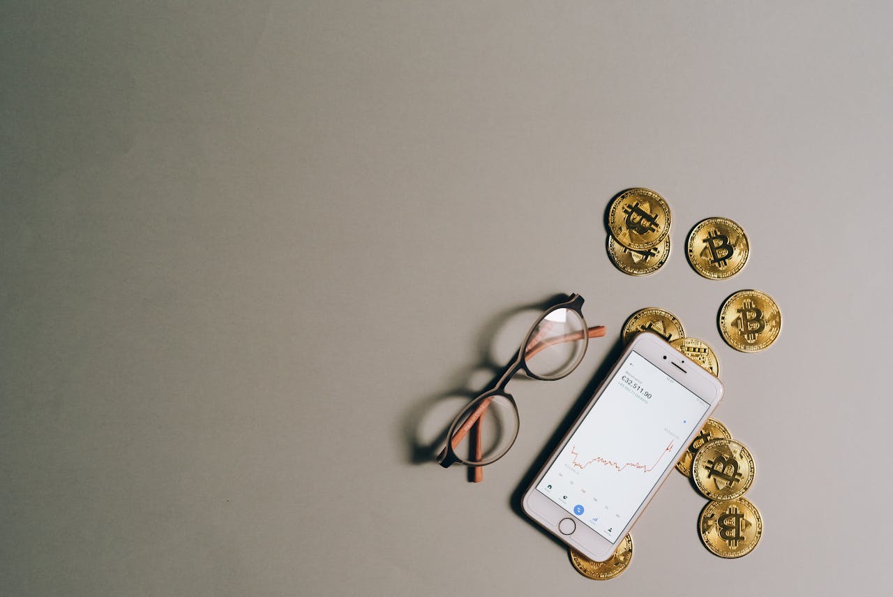hero-img-02 Flat lay of Bitcoin coins, smartphone with graph, and glasses on gray surface.